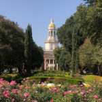 View of a college campus with a red building topped with a tall, white rotunda sits behind tall green trees, with pink rose bushes in the foreground