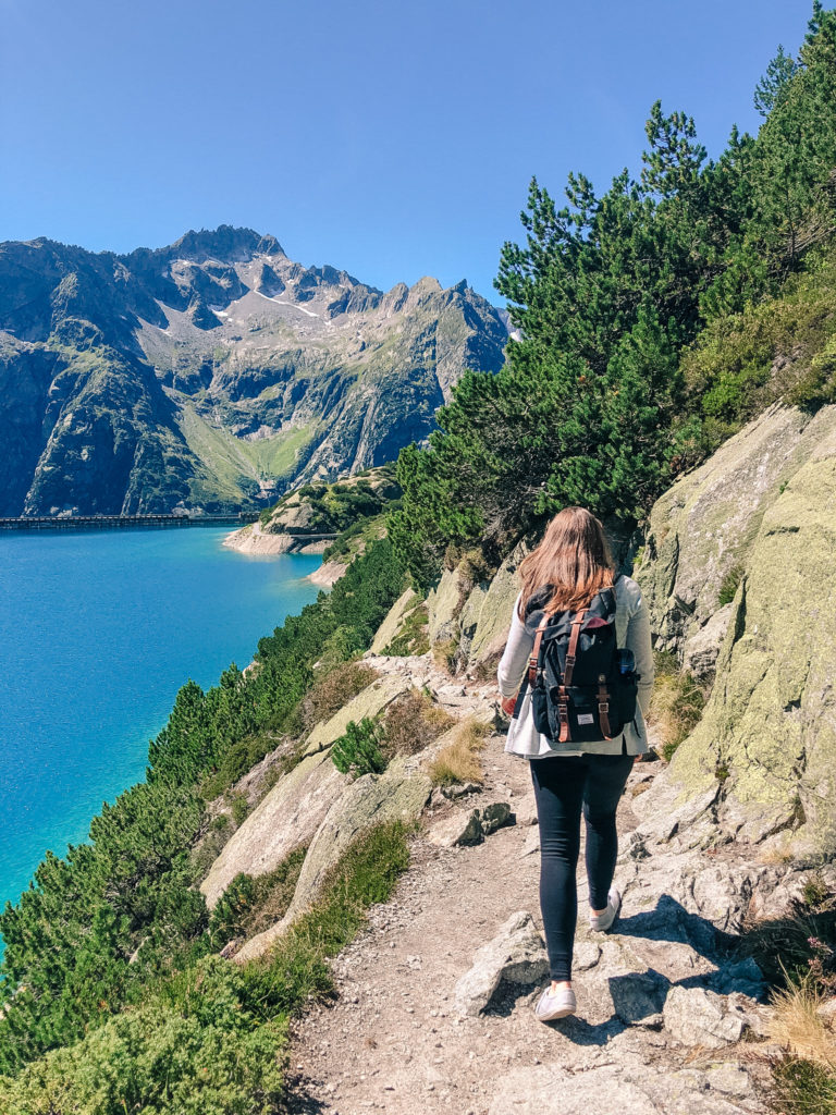 Girl wearing black tights, a grey sweater, and a black backpack walks away from the camera on a rocky, dirt path with a stone mountain wall on the right side and a bright blue lake surrounded by short, green plants on the left side