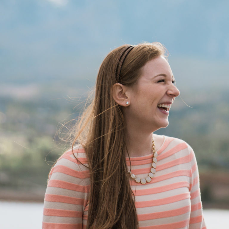Girl with long, brown hair wearing an orange and cream striped shirt looks off to the right side of the camera laughing, with a blurred lake in the background