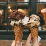 Two hands holding sugar cones topped with vanilla and chocolate ice cream (on the left) and caramel ice cream (on the right) with a blurred store window in the background