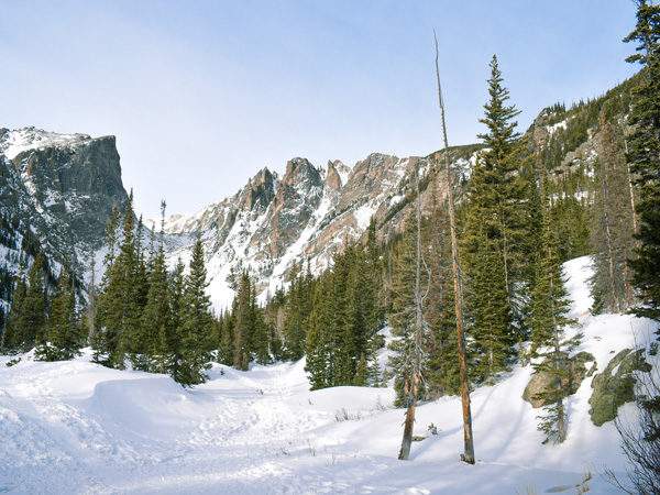 Deep snow-covered meadow in front of mountain peaks forming a "U" shape, with green pine trees at the base of the mountain