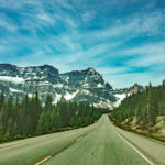 Long straight paved road lined with tall, green pine trees, looking towards craggy, grey mountains with bands of snow covering them