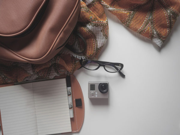 White table laid out with a brown backpack, white notebook, small camera, pair of glasses, and an orange scarf