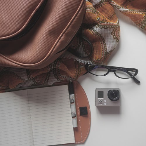 White table laid out with a brown backpack, white notebook, small camera, pair of glasses, and an orange scarf