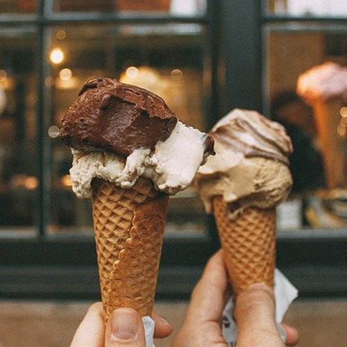 Two hands holding sugar cones topped with vanilla and chocolate ice cream (on the left) and caramel ice cream (on the right) with a blurred store window in the background