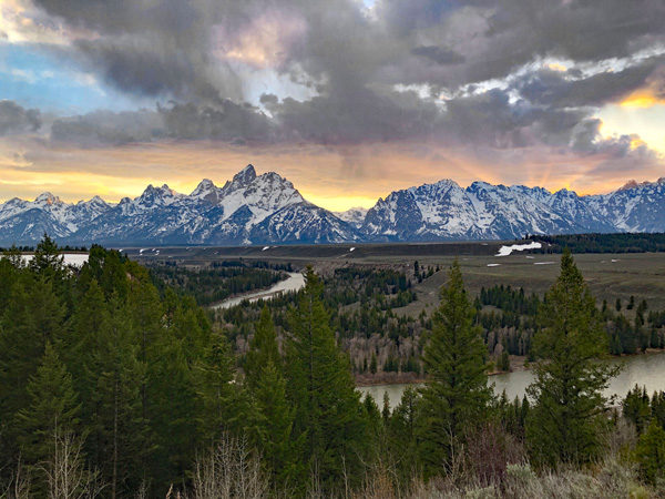 Panoramic view of jagged mountains covered in snow in the background, with a winding river and tall pine trees in the foreground, as the sun sets amid grey clouds behind the mountains