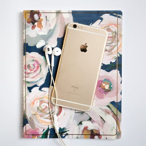 Navy, pink, gold, and white floral-printed notebook sits on a white surface, with a gold iPhone and a pair of white headphones sitting on top of the notebook