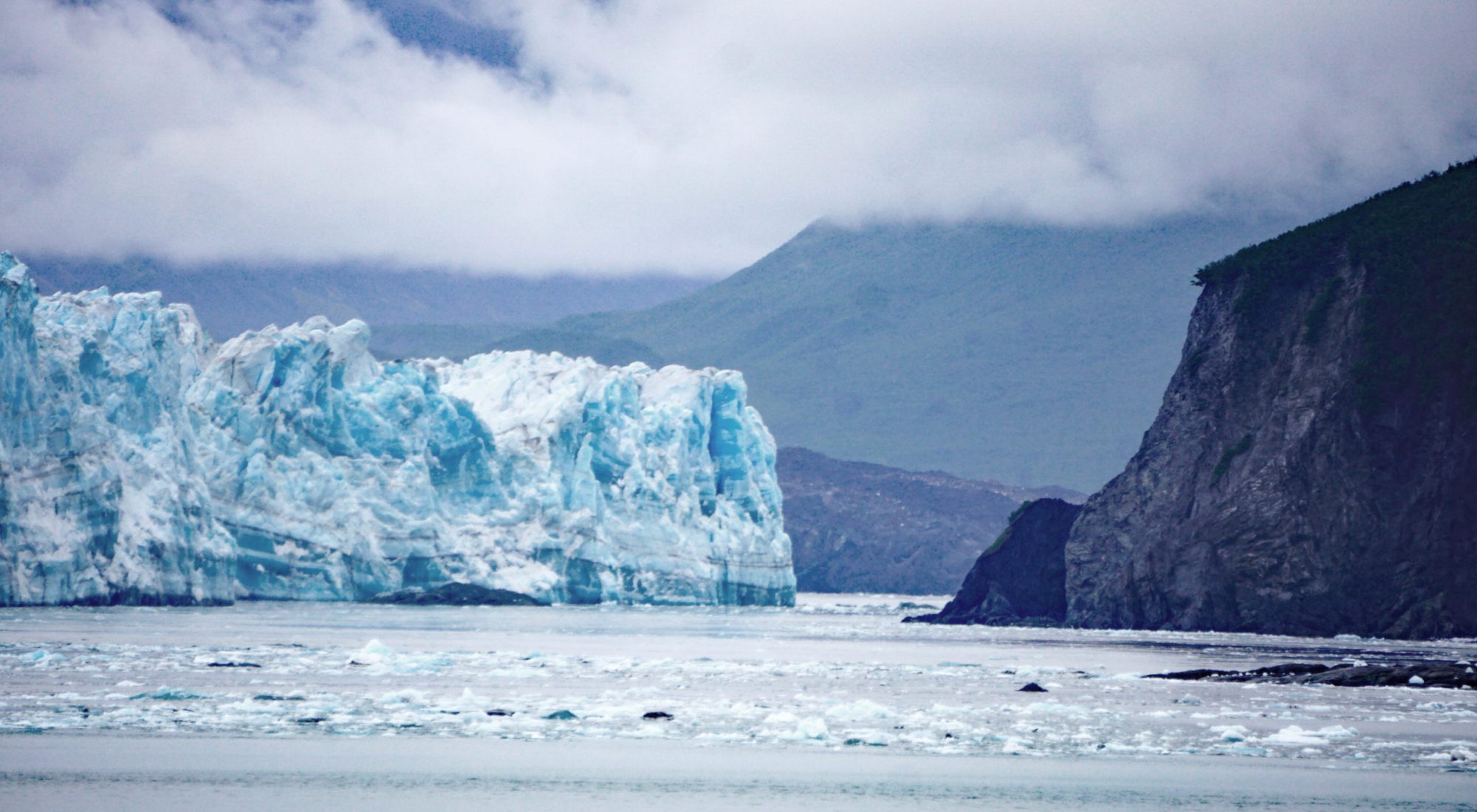 Ice-lined grey water with a large white and blue glacier on the left and a dark grey mountain on the right, with a hazy green hill and grey, cloud-covered sky in the background