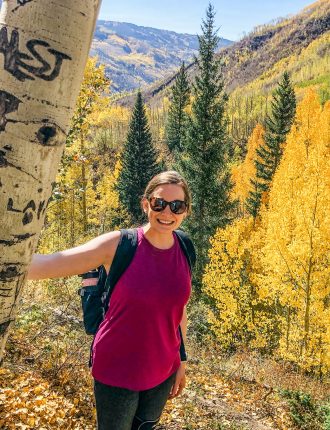 Smiling girl in purple tank top and grey tights wearing a black backpack poses next to an aspen tree, surrounded by bright yellow aspens and a couple of dark green pine trees